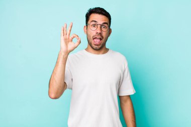 young handsome hicpanic man feeling successful and satisfied, smiling with mouth wide open, making okay sign with hand