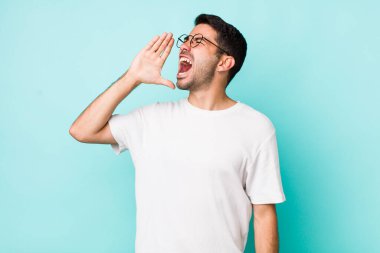 young handsome hicpanic man yelling loudly and angrily to copy space on the side, with hand next to mouth