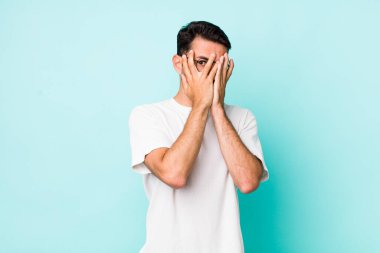 young handsome hicpanic man feeling scared or embarrassed, peeking or spying with eyes half-covered with hands