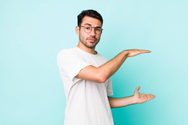 young handsome hicpanic man holding an object with both hands on side copy space, showing, offering or advertising an object