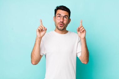 young handsome hicpanic man feeling awed and open mouthed pointing upwards with a shocked and surprised look