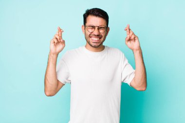 young handsome hicpanic man smiling and anxiously crossing both fingers, feeling worried and wishing or hoping for good luck