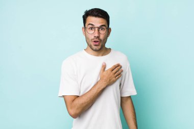 young handsome hicpanic man feeling shocked and surprised, smiling, taking hand to heart, happy to be the one or showing gratitude
