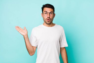 young handsome hicpanic man looking surprised and shocked, with jaw dropped holding an object with an open hand on the side