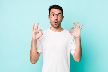 young handsome hicpanic man feeling shocked, amazed and surprised, showing approval making okay sign with both hands