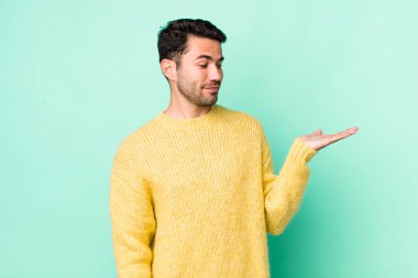 young handsome hicpanic man feeling happy and smiling casually, looking to an object or concept held on the hand on the side