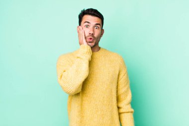 young handsome hicpanic man feeling shocked and astonished holding face to hand in disbelief with mouth wide open