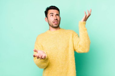 young handsome hicpanic man shrugging with a dumb, crazy, confused, puzzled expression, feeling annoyed and clueless