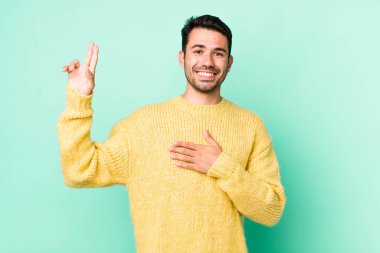 young handsome hicpanic man looking happy, confident and trustworthy, smiling and showing victory sign, with a positive attitude