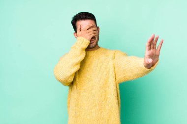 young handsome hicpanic man covering face with hand and putting other hand up front to stop camera, refusing photos or pictures