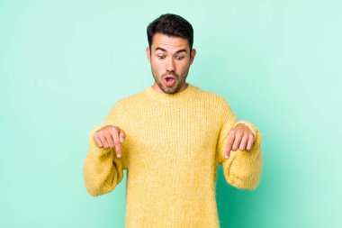 young handsome hicpanic man feeling shocked, open-mouthed and amazed, looking and pointing downwards in disbelief and surprise
