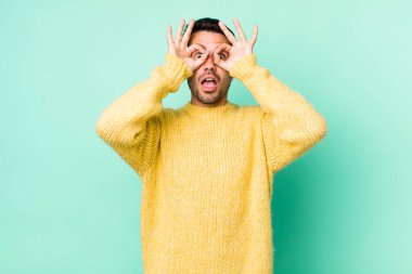 young handsome hicpanic man feeling shocked, amazed and surprised, holding glasses with astonished, disbelieving look