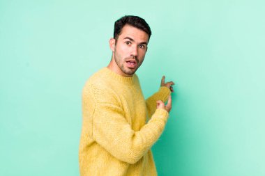 young handsome hicpanic man feeling shocked and surprised, pointing to copy space on the side with amazed, open-mouthed look