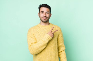 young handsome hicpanic man feeling happy, positive and successful, with hand making v shape over chest, showing victory or peace