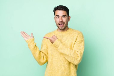 young handsome hicpanic man smiling cheerfully and pointing to copy space on palm on the side, showing or advertising an object
