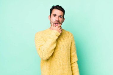 young handsome hicpanic man with mouth and eyes wide open and hand on chin, feeling unpleasantly shocked, saying what or wow