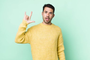 young handsome hicpanic man feeling happy, fun, confident, positive and rebellious, making rock or heavy metal sign with hand