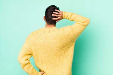 young handsome hicpanic man feeling clueless and confused, thinking a solution, with hand on hip and other on head, rear view