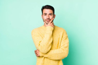 young handsome hicpanic man looking serious, thoughtful and distrustful, with one arm crossed and hand on chin, weighting options