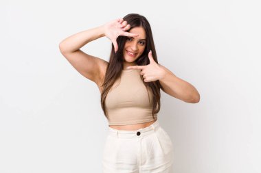 Genç ve güzel bir kadın mutlu, arkadaş canlısı ve pozitif hissediyor, gülümsüyor ve elleriyle bir portre veya fotoğraf çerçevesi yapıyor.