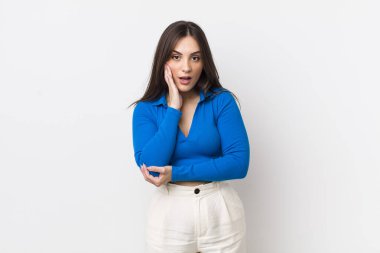 Genç ve güzel bir kadın şok ve inançsızlık içinde, elleri yanağında ve kolları çaprazlanmış, sersemlemiş ve şaşırmış hissediyor.