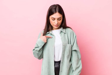 Genç ve güzel bir kadın neşeyle gülümsüyor, yere bakıyor ve göğsüne işaret ediyor.