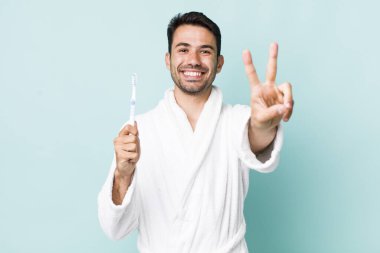 young adult hispanic man smiling and looking happy, gesturing victory or peace. toothbrush concept