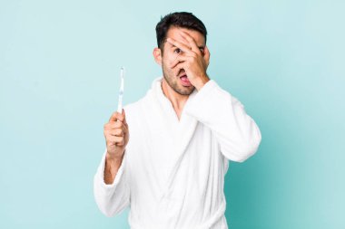 young adult hispanic man looking shocked, scared or terrified, covering face with hand. toothbrush concept