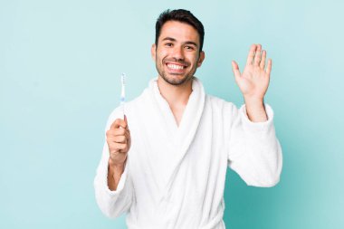 young adult hispanic man smiling happily, waving hand, welcoming and greeting you. toothbrush concept