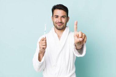 young adult hispanic man smiling and looking friendly, showing number one. toothbrush concept