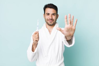 young adult hispanic man smiling and looking friendly, showing number five. toothbrush concept