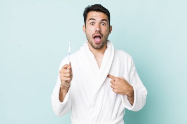 young adult hispanic man looking shocked and surprised with mouth wide open, pointing to self. toothbrush concept