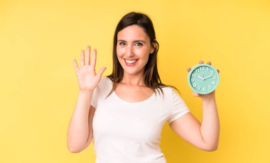 Genç ve güzel bir kadın mutlu bir şekilde gülümsüyor, el sallıyor, sizi selamlıyor ve selamlıyor. Alarm saati kavramı
