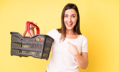 Genç ve güzel bir kadın şok olmuş ve ağzı açık bir şekilde kendini işaret ederek şaşırmış görünüyor. Alışveriş sepeti konsepti