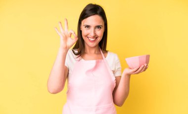 Genç ve güzel bir kadın kendini mutlu hissediyor ve onayını veriyor. Ev aşçısı konseptinde yemek pişirmek