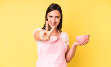 Genç ve güzel bir kadın gülümsüyor ve mutlu görünüyor. Zafer ya da barış işareti yapıyor. Ev aşçısı konseptinde yemek pişirmek