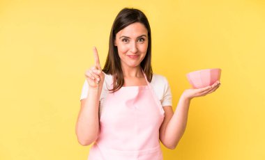 Genç ve güzel bir kadın gülümsüyor ve arkadaş canlısı görünüyor. Bir numarayı gösteriyor. Ev aşçısı konseptinde yemek pişirmek