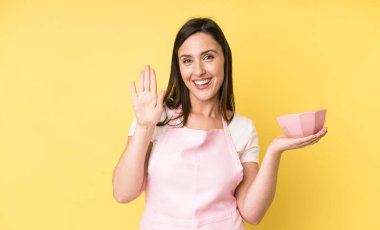 Genç ve güzel bir kadın mutlu bir şekilde gülümsüyor, el sallıyor, sizi selamlıyor ve selamlıyor. Ev aşçısı konseptinde yemek pişirmek