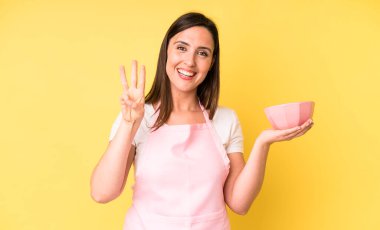 Genç ve güzel bir bayan gülümsüyor ve arkadaş canlısı görünüyor, üç numarayı gösteriyor. Ev aşçısı konseptinde yemek pişirmek