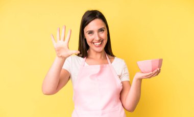 Genç ve güzel bir kadın gülümsüyor ve arkadaş canlısı görünüyor. Beş numarayı gösteriyor. Ev aşçısı konseptinde yemek pişirmek