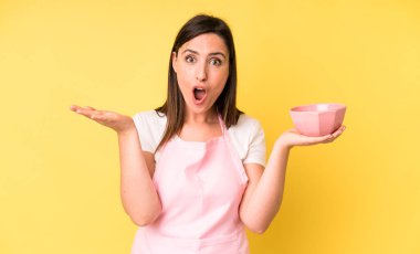 Genç ve güzel bir kadın şaşırmış ve şok olmuş görünüyor. Çenesi düşmüş ve elinde bir nesne tutuyor. Ev aşçısı konseptinde yemek pişirmek