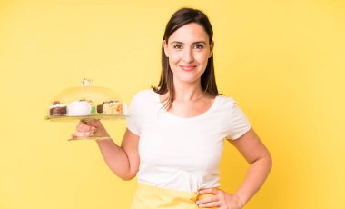 Genç ve güzel bir kadın mutlu bir şekilde gülümsüyor, eli kalçasında ve kendinden emin. Ev yapımı kek konsepti