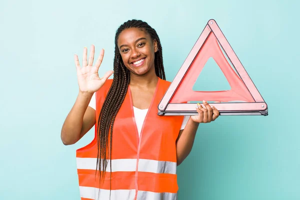 young adult black woman smiling and looking friendly, showing number five. car emergency triangle concept