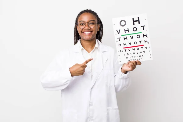 young adult black woman smiling cheerfully, feeling happy and pointing to the side. optical vision test concept