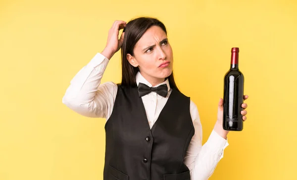 young adult pretty woman feeling puzzled and confused, scratching head. waiter and wine bottle concept