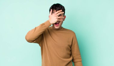 Genç, Latin bir adam şok olmuş, korkmuş ya da korkmuş görünüyor. Yüzü elle kaplanmış ve parmaklarının arasından gizlice bakıyor.