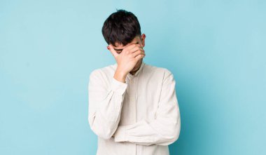 Genç, İspanyol asıllı bir adam stresli, utanmış ya da üzgün görünüyor, baş ağrısı var, yüzünü eliyle kapatıyor.