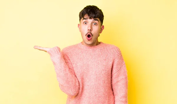 stock image young adult hispanic man looking surprised and shocked, with jaw dropped holding an object with an open hand on the side