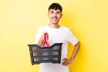 Genç İspanyol adam mutlu bir şekilde gülümsüyor, eli kalçasında ve kendinden emin. boş alışveriş sepeti kavramı