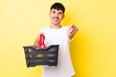 Genç İspanyol adam kamerayı göstererek seni seçiyor. boş alışveriş sepeti kavramı